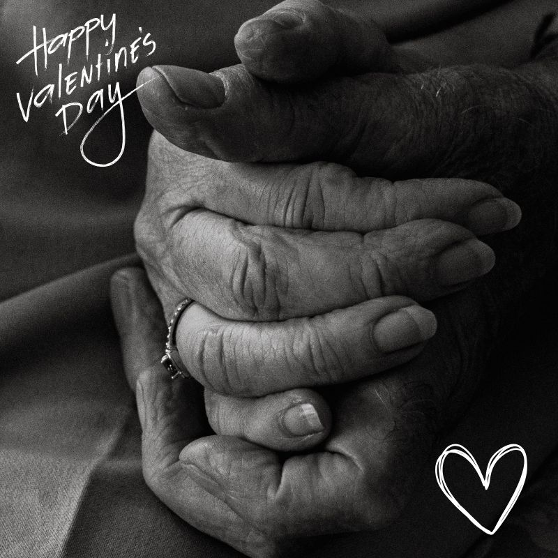 Elderly couple holding hands with ‘Happy Valentine’s Day’ written in cursive, symbolizing love, caregiving, and devotion on Valentine’s Day.