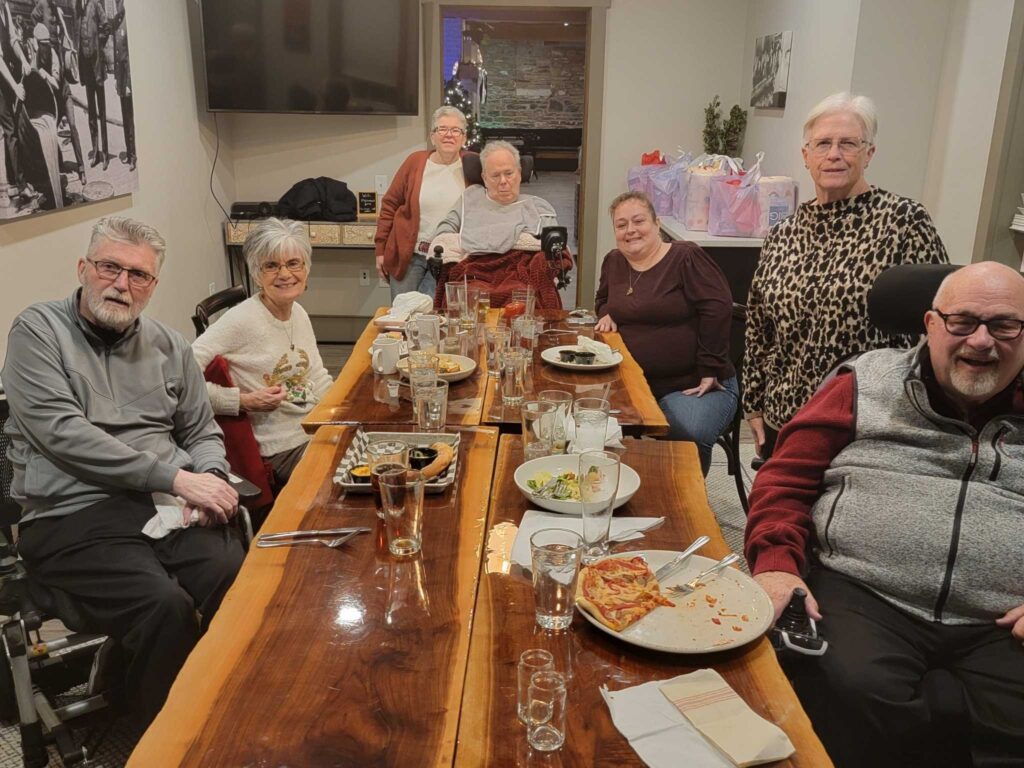 ALS patients and caregivers gathered for a community dinner, sharing support, stories, and connection around a long wooden table.