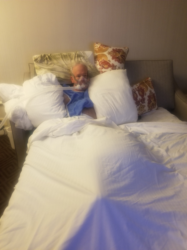 Man with ALS using a ventilator, surrounded by pillows in a hotel bed too high for safe transfers, showing real challenges behind so-called accessible rooms