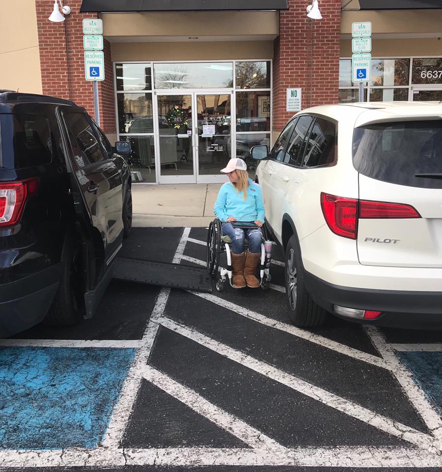 Woman using a wheelchair blocked between two vehicles in a Van Accessible parking space, illustrating lack of practical accessibility during Disability Pride Month