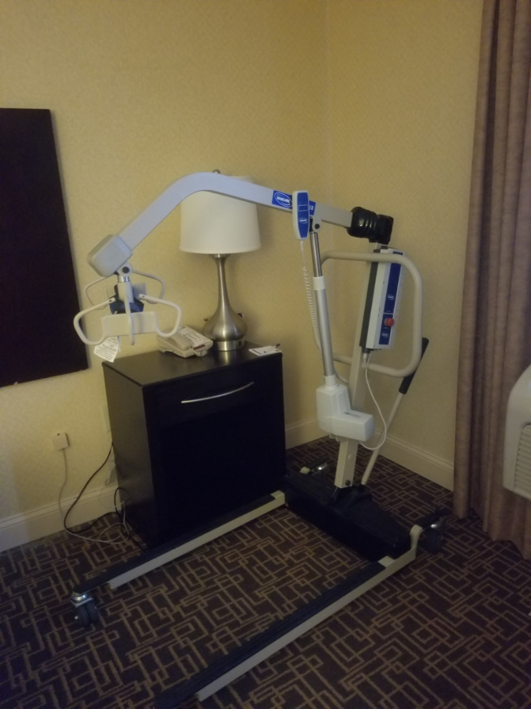 Medical patient lift next to hotel nightstand, used for transferring individuals with ALS or mobility disabilities who cannot stand or move independently